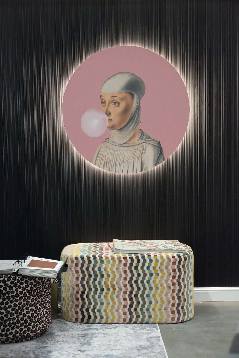 Portret kobiety w stylu renesansowym z bąblem gumy do żucia, umieszczony w okrągłym, oświetlonym otworze na ciemnej, pionowo prążkowanej ścianie, nad kolorowym, wzorzystym pufem i koszem w panterkę.