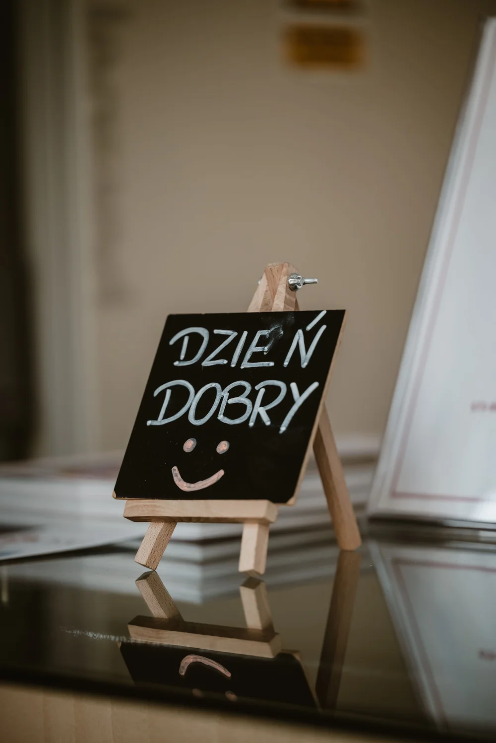 Mała tabliczka kredowa na drewnianym statywie z napisem „DZIEŃ DOBRY” i uśmiechniętą buźką, umieszczona na lśniącym, szklanym blacie.
