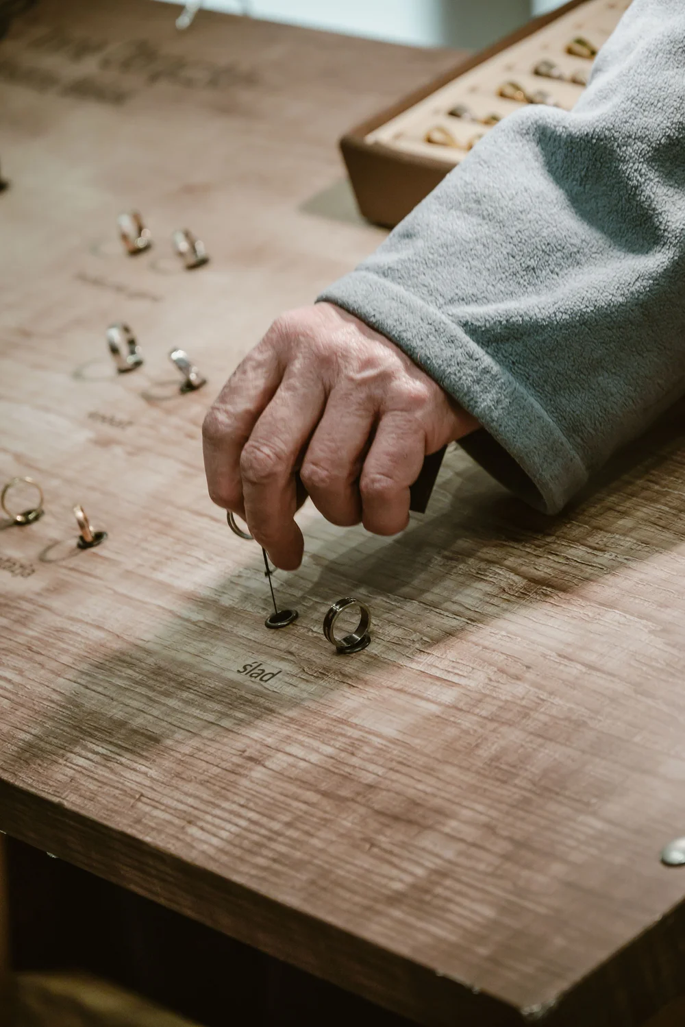 Ręka w szarej bluzie dotyka pierścionka na drewnianym stole z ułożonymi w rzędach innymi obrączkami i napisami.