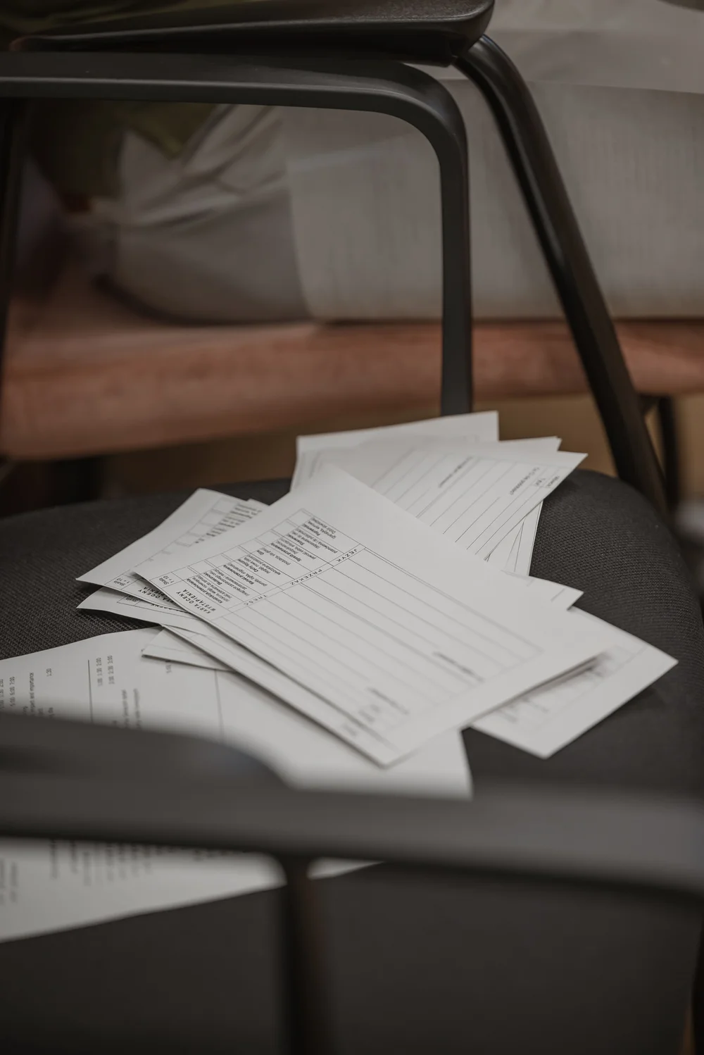 Stos papierów z drukowanymi formularzami leżący na siedzisku czarnego krzesła, w tle widoczne nogi drugiego krzesła i fragment mebla.