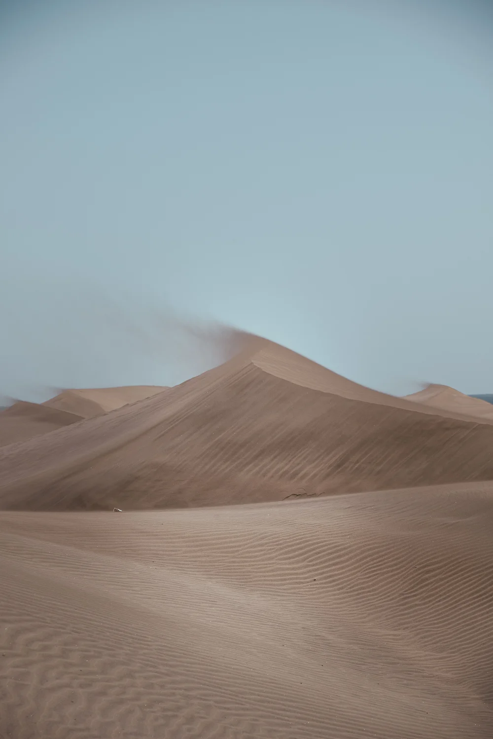 Pustynne wydmy z delikatnymi liniami piasku, pod jasnoniebieskim niebem, z lekkim wiatrem unoszącym piasek w tle.