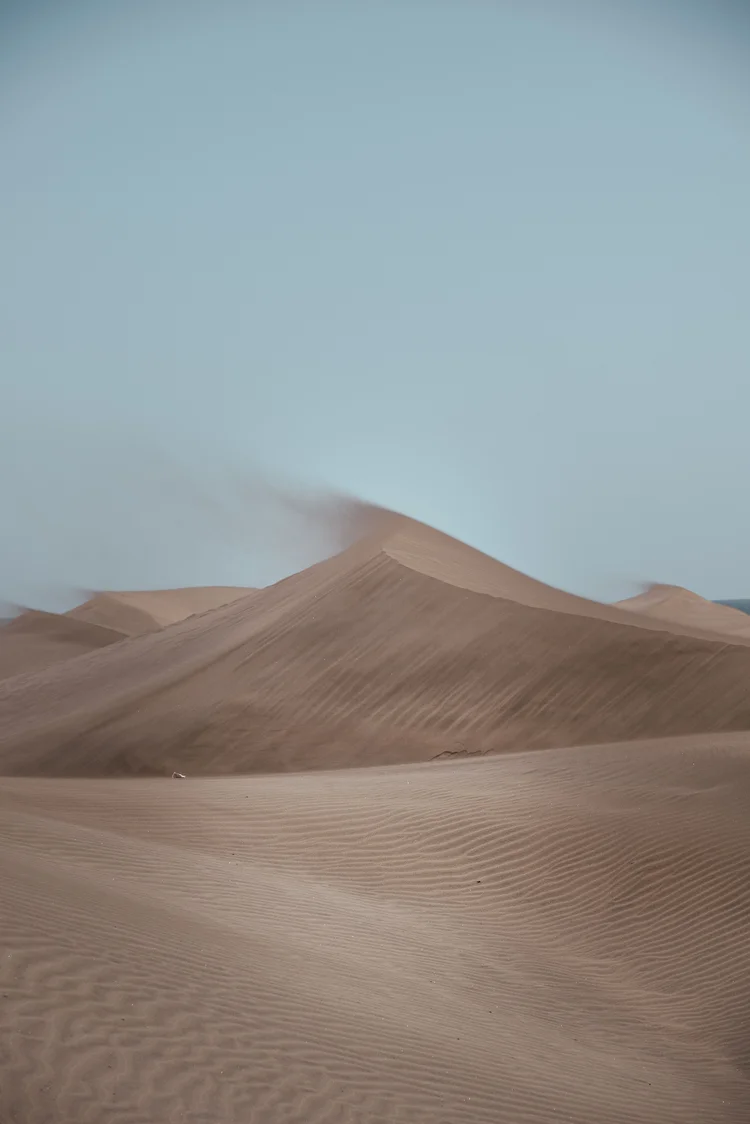 Pustynne wydmy z delikatnymi liniami piasku, pod jasnoniebieskim niebem, z lekkim wiatrem unoszącym piasek w tle.