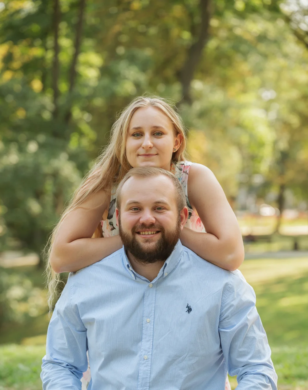Mężczyzna w niebieskiej koszuli z brodą, uśmiechnięty, niesie na plecach młodą kobietę o długich blond włosach, ubraną w kwiecistą sukienkę, na tle zielonego parku.
