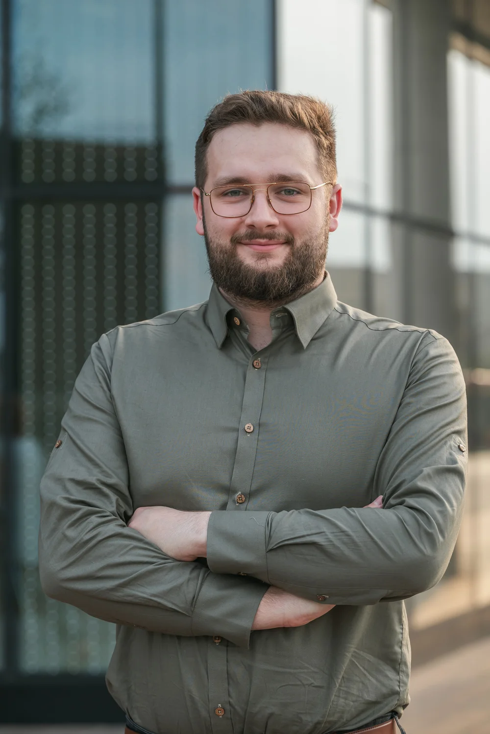 Mężczyzna z brodą i okularami w zielonej koszuli, uśmiechnięty, z rękami skrzyżowanymi na piersi, stojący przed szklanym budynkiem.