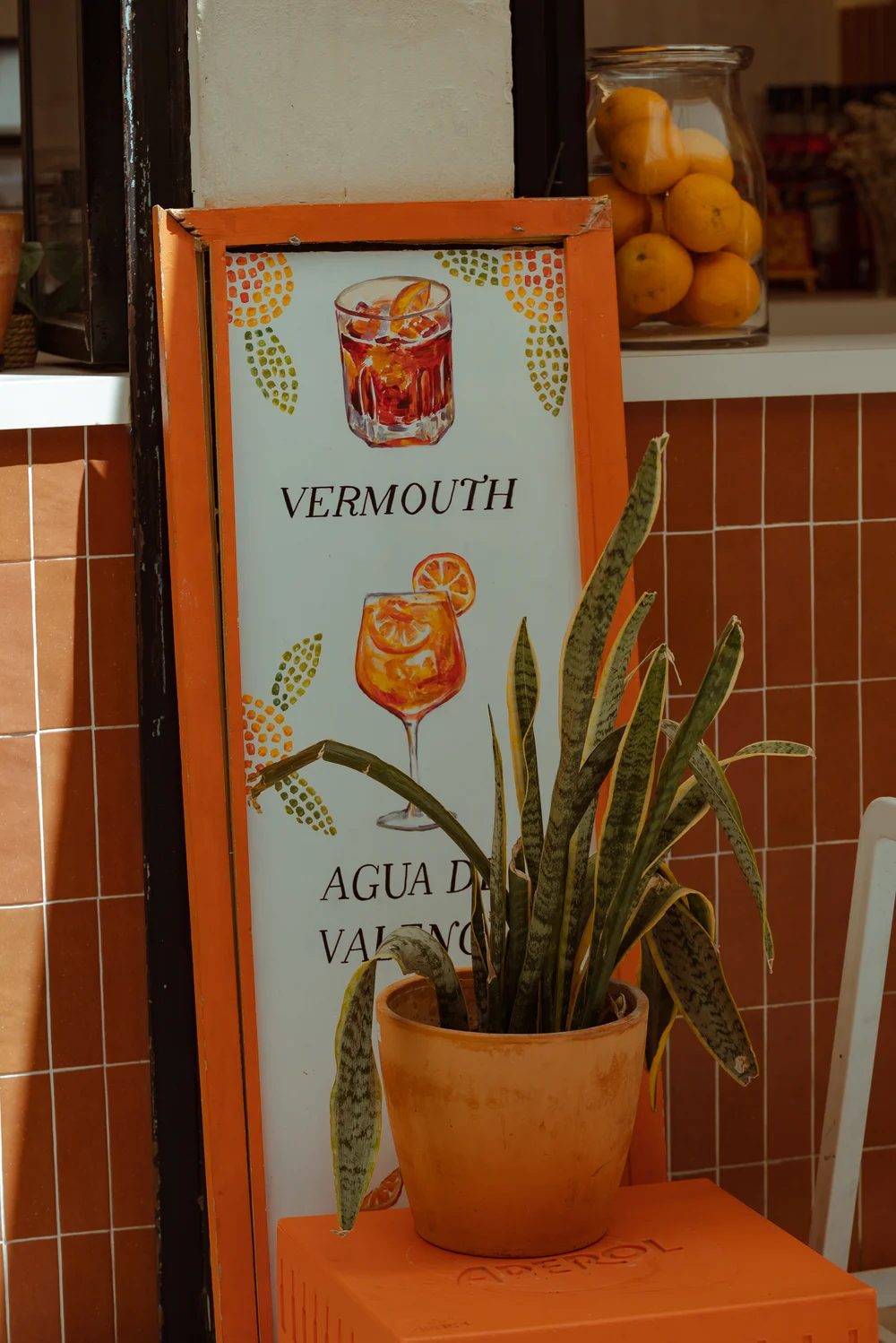 Oznaka z ilustracjami drinków: Vermouth i Agua de Valencia, ozdobiona pomarańczami i rośliną w doniczce.